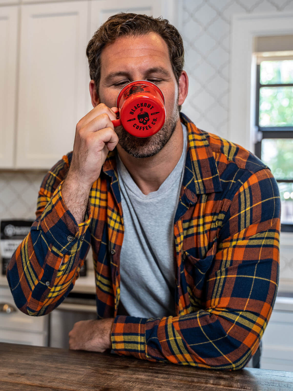 Man in a plaid shirt drinking from a red mug in a kitchen.