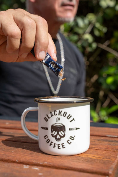 Person adding a coffee pod to a 'Blackout Coffee Co.' mug outdoors.