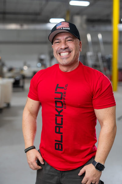 Man wearing a red 'Blackout' t-shirt and black cap with an American flag in a warehouse setting.