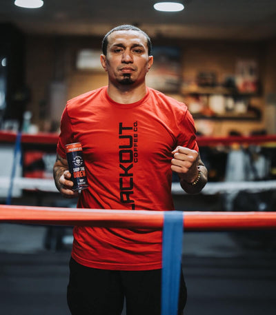 Man in a red 'BLACKOUT' shirt holding a supplement bottle in a boxing ring.