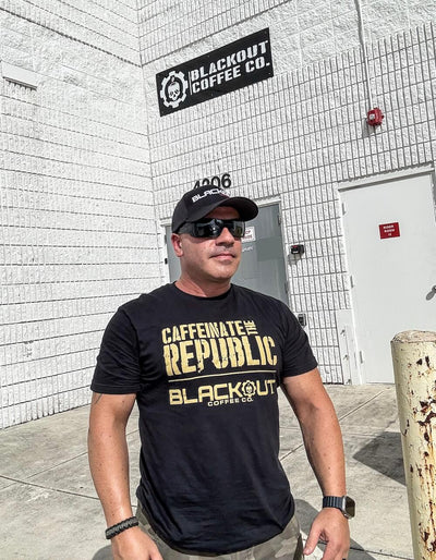John Santos wearing a black 'Blackout Coffee Co.' t-shirt standing in front of a building with the same branding.