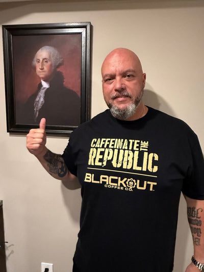 Man wearing a black t-shirt with 'Caffeinate the Republic Blackout Coffee Co.' text, giving a thumbs-up in front of a portrait on a wall.