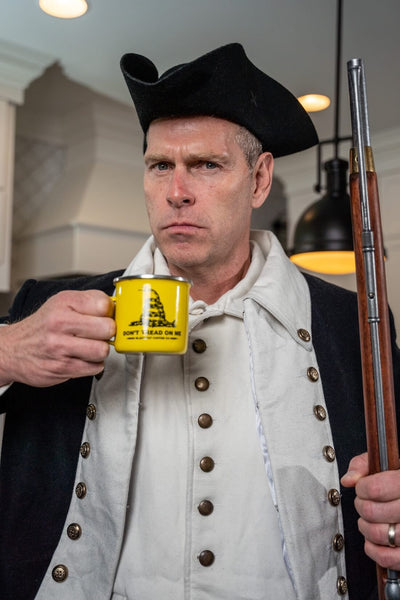 Man in colonial attire holding a rifle and a mug with a flag design.