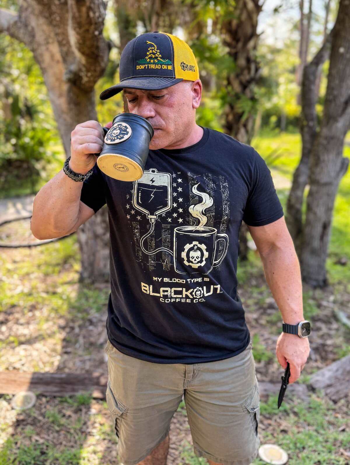John Santos drinking from a coffee cup outdoors, wearing a black t-shirt with graphics and text.