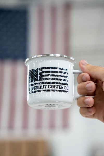 Hand holding a mug with an American flag design and 'Lockout Coffee Co.' text, blurred flag in the background.
