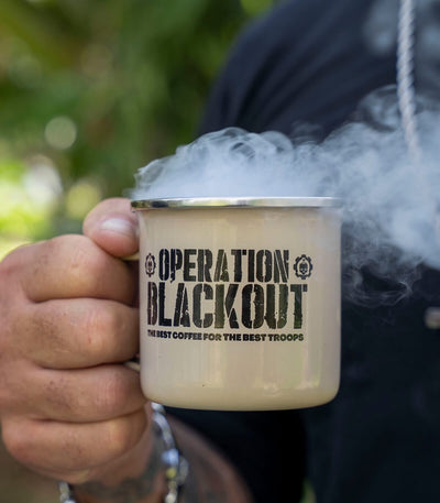 Person holding a 'Operation Blackout' coffee mug with steam, outdoor setting