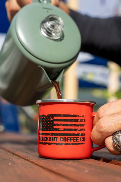Green thermos pouring coffee into a red 'Blackout Coffee Co.' mug on a wooden surface.